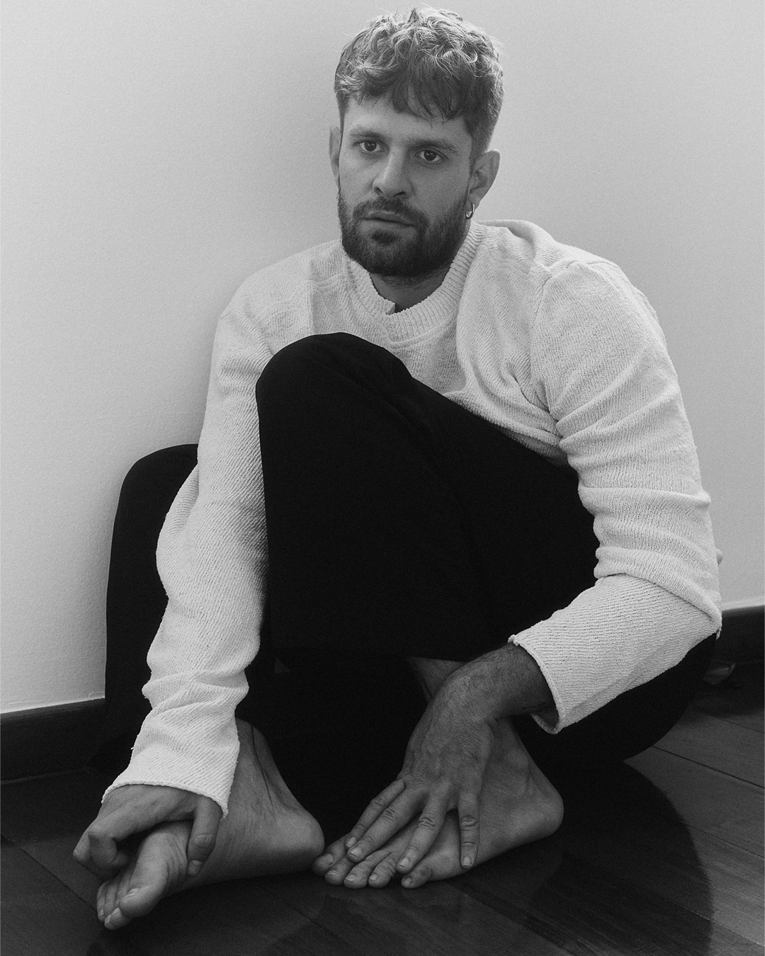 Ator Alejandro Claveaux sentado no chão, de calça preta e camisa branca.