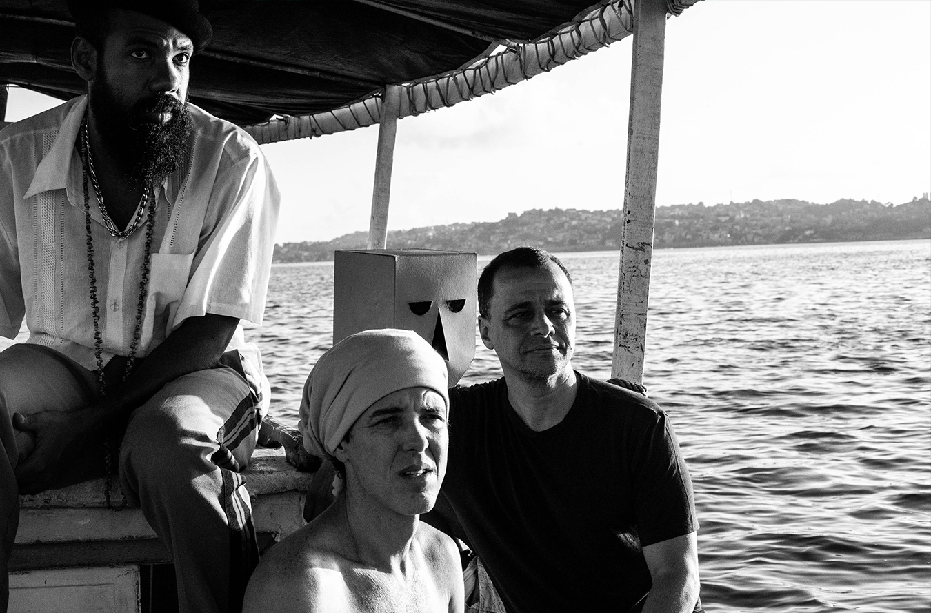 Todos os integrantes da banda em um barco, atrás é possível ver o mar e vegetação.